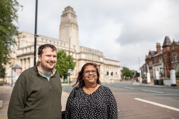 Two mature students stood at a roadside smiling. The University's Parkinson Building is in the background.