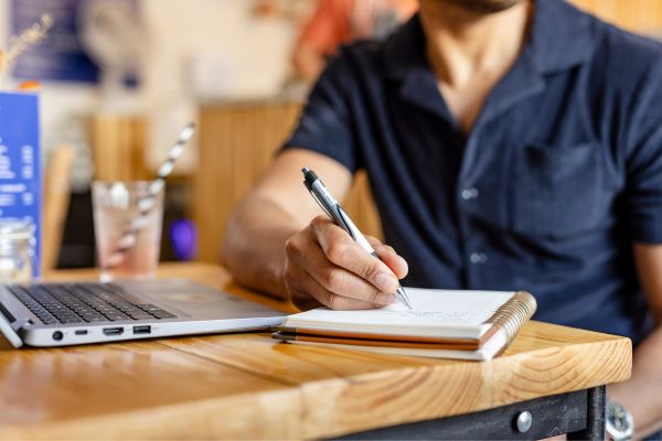 A hand taking notes on on a notepad, with a laptop open on the table.