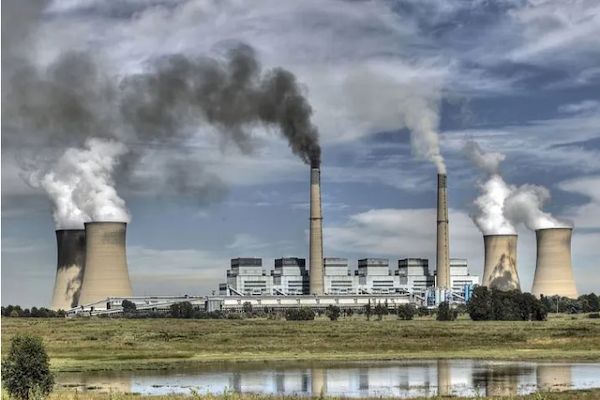 Barren landscape with large power station in middle; with multiple short and long chimneys emitting smoke against a blue sky