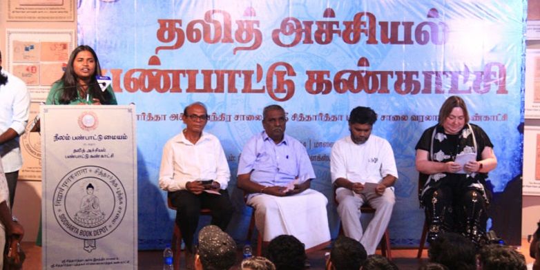 A group of people sat on a stage. Next to them, another person is stood speaking at a lectern.