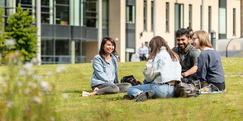 students sat on the grass on campus on a sunny day, chatting and smiling