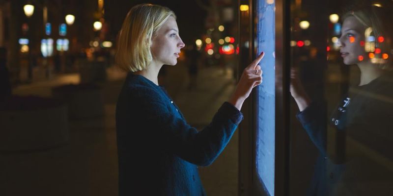A woman stands in a dark street at night pressing a touch screen on a board.