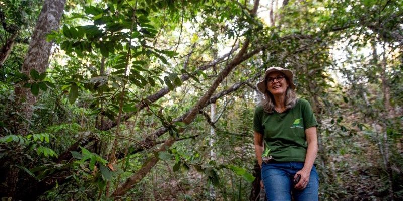 Professor Beatriz Schwantes Marimon stands in the thick of the Amazon rainforest, with entangled trees growing behind her. She is smiling and is wearing a hat, a t-shirt and jeans.