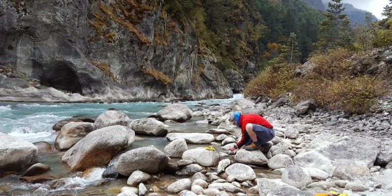 A researcher kneels on rocks next to a river in a valley, studying the river and rocks below.