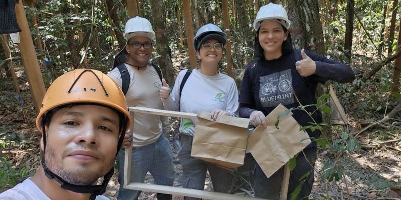 Carlos Dias, Eder Carvalho, Melina Arantes and Carla Luz stand in a group, taking a selfie. They are wearing hard hats and three are holding a large wooden frame. They smile at the camera and Carla holds her thumb up.