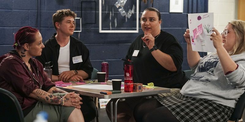 Four people sat around a table, having a discussion. One of them is holding up a piece of paper with notes on it.