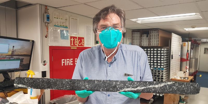 Dr Andrew McCaig holding a long core section of Harzburgite rock from the Earth’s mantle. he is wearing a mask and gloves as a precaution