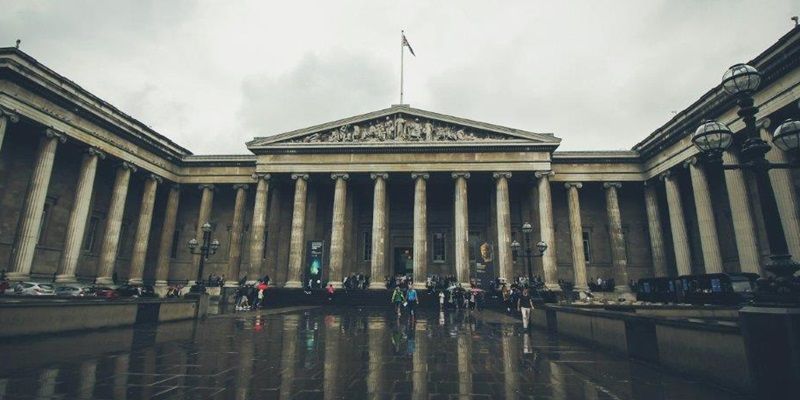 The imposing columns and forecourt of the British Museum on a rainy day with people outside the building.
