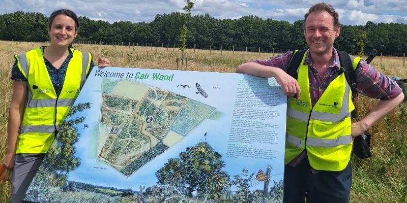 Cat and Tom, the academic leads for Gair Wood, lean on signage displaying the different areas of the wood.