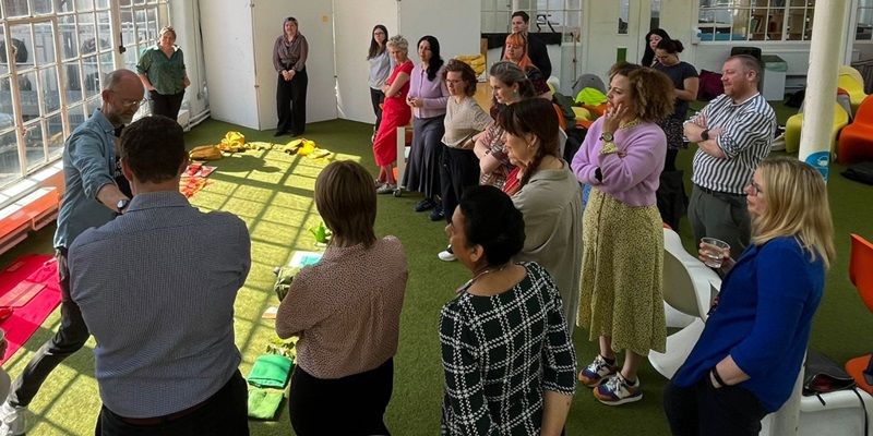 A group of around twenty people stood around, taking part in a workshop and listening to a facilitator demonstrating an activity.