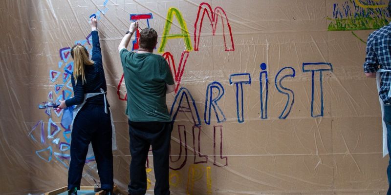 Two people, their backs to camera, painting words and pictures on a very large canvas.