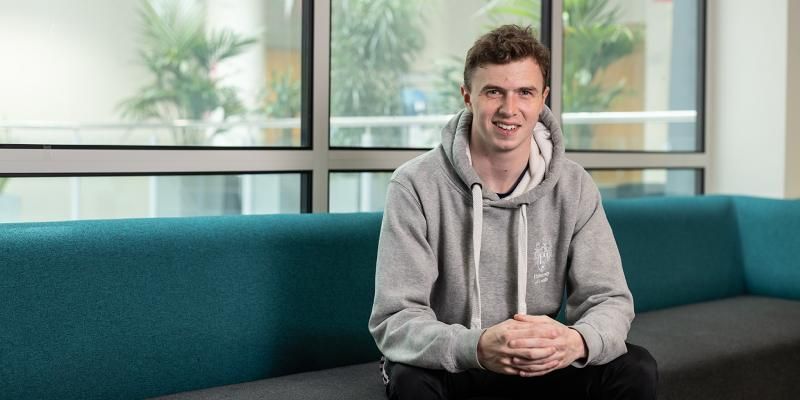 A student wearing a grey University of Leeds hoodie sitting with their hands together on a sofa