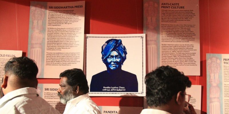 People walking past a wall covered with posters. One says 'Sri Siddartha Press', another says' Anti-Caste print Culture.' Another features a head and shoulders graphic print of a person wearing a turban.