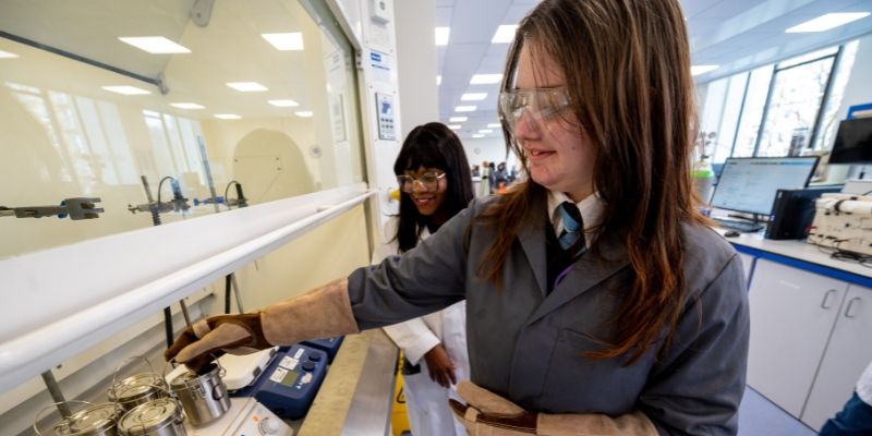 High school pupil, wearing lab coat and protective gloves, places small metal canisters onto a piece of equipment in a lab. They are supervised by a person in a white lab coat.