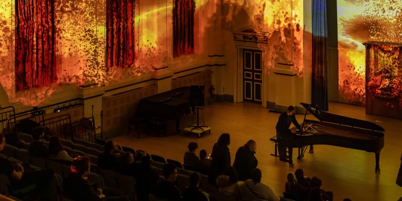 A concert hall is lit up with a fiery red and orange projection. On the stage floor, someone stands over a piano. People in the audience sit, watching the performance.