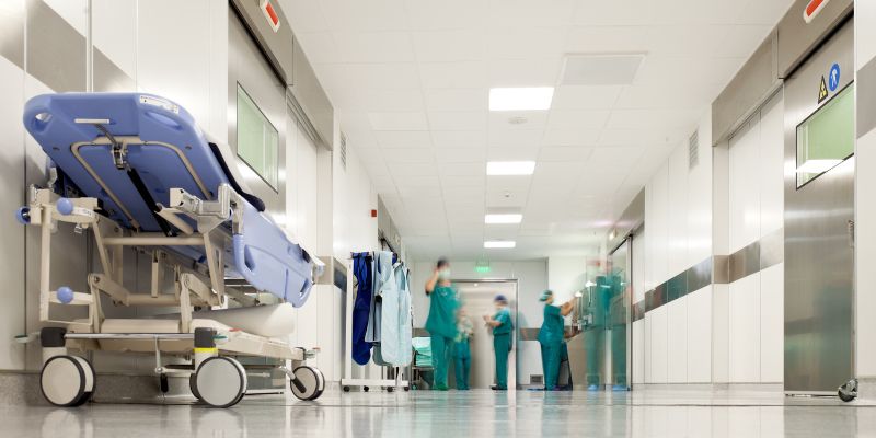 A hospital corridor with an empty bed to the left and medical workers in the background.