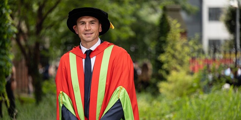 Kevin Sinfield stood outside on campus on a sunny day wearing graduation cap and gown.