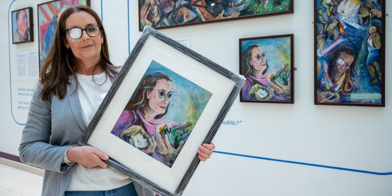 Trial participant Rebecca holding her painting in front of the I'm Still Me exhibition in the Bexley Wing at St James' University Hospital.