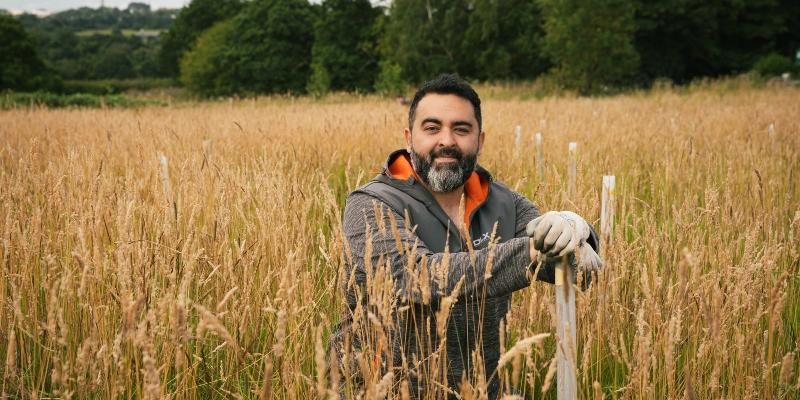 Nader Mabadi crouching next to a plastic tree guard in a field of long grass and planted trees.