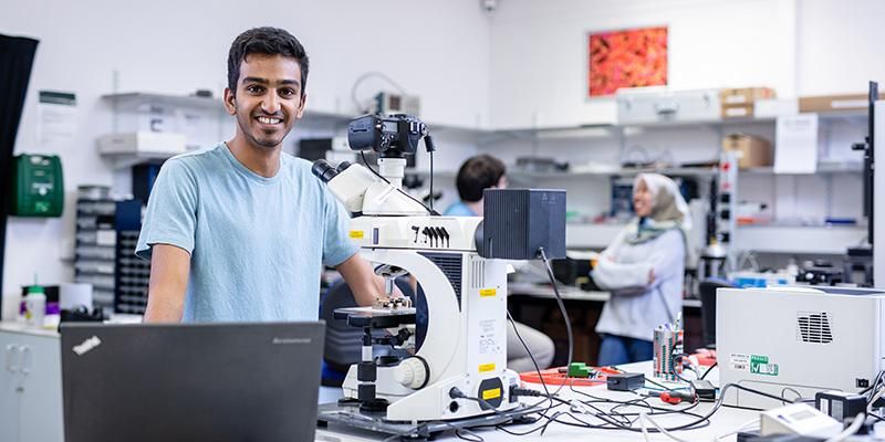 A busy lab with lots of equipment and a postgraduate researcher smiling with a laptop