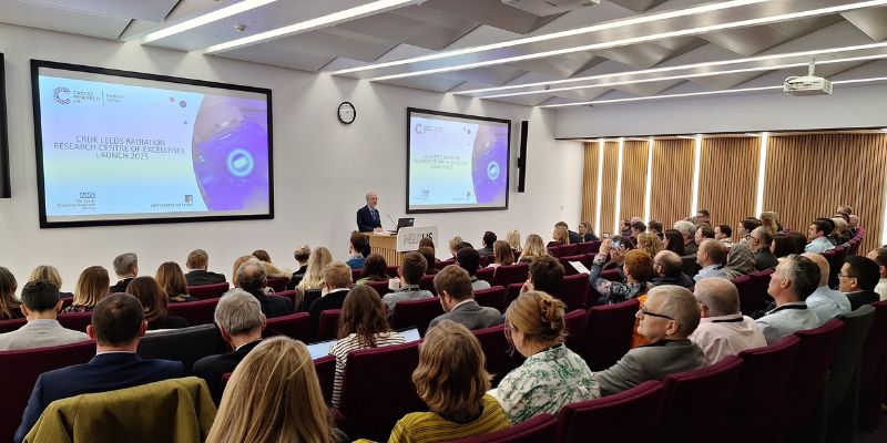 A crowd in a modern lecture theatre listens to a talk by a professor. Two screens display logos for CRUK RadNet Leeds.