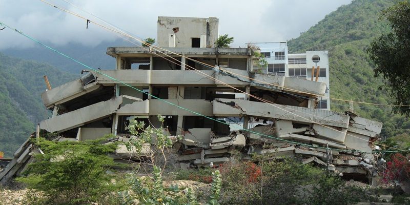 A large concrete building which has collapsed, with mountains in the background.