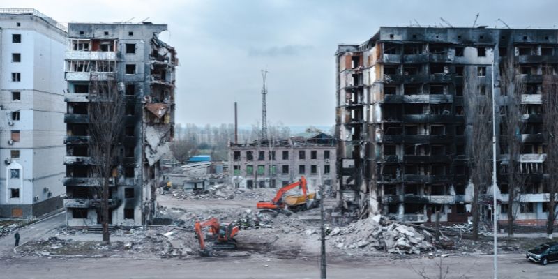 Fire-blackened multi-storey buildings destroyed by war in Ukraine, stand in rubble, beside diggers.