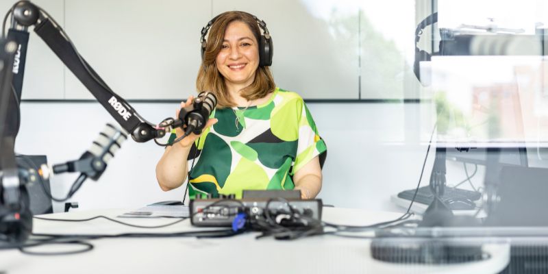 A tutor, smiling, sits at a table with recording equipment set up in front of them. They are wearing headphones.