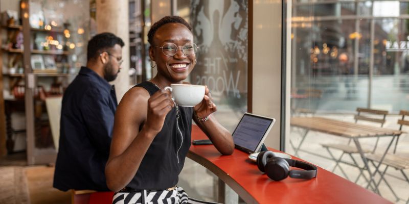 A student sat in a cafe, with a tablet and headphones on the table in front of them. They are holding a cup of coffee and smiling.