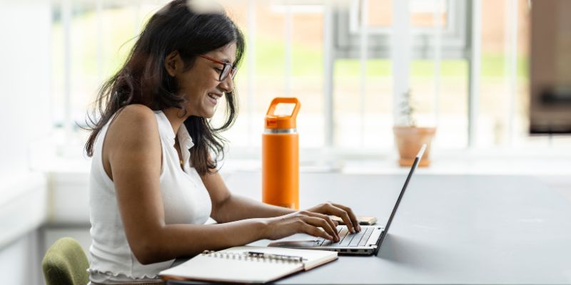 A student working on their laptop at a table, smiling at the screen.