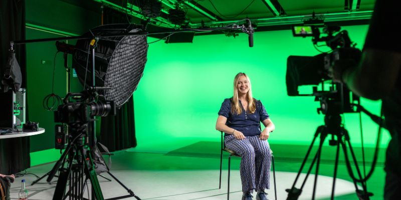 A University course tutor sitting on a chair, taking part in filming. There are cameras set up and a green screen.