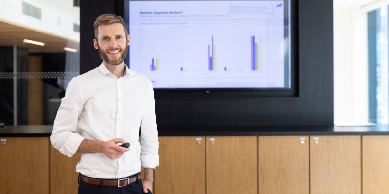 A person delivering a presentation. They are standing in front of a screen showing bar charts, holding a remote control.