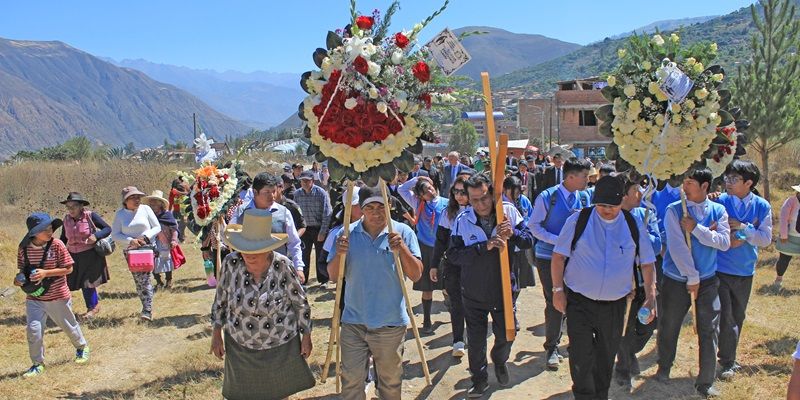 A large memorial procession of people carrying flower garlands and a wooden cross up a hillside. There are mountains in the background.