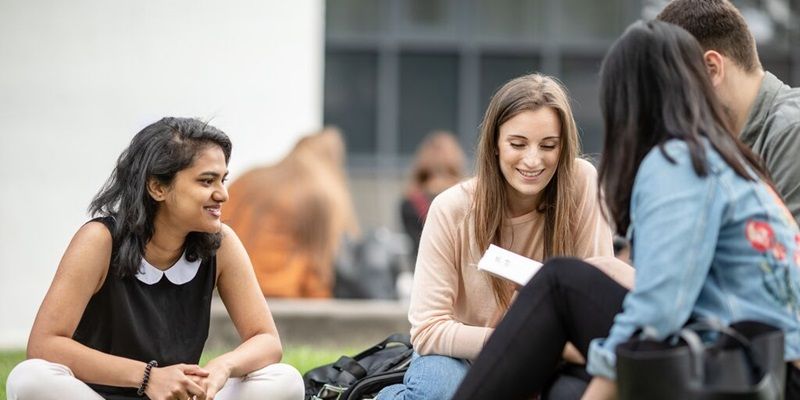 Four students sat outside on campus in summer. One is holding a book or document and they are chatting.