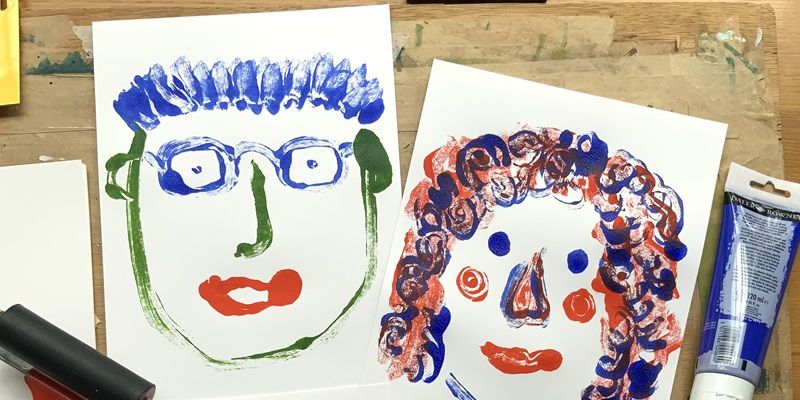 Two monoprints of drawings of faces on a table with a small inking roller and tube of paint