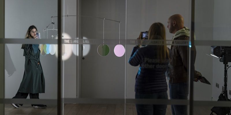 Three people looking at and photographing a hanging sculpture of coloured discs.