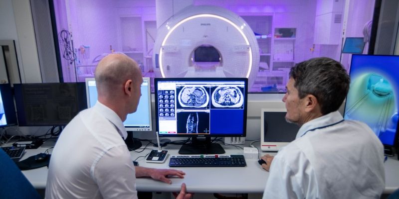 Two researchers looking at a scan behind a glass window in the radiotherapy department at St James' Hospital, Leeds. On the other side of the window is the MR Simulator machine 