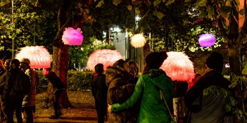 Multisensory artwork is hung from trees with visitors looking at the art.