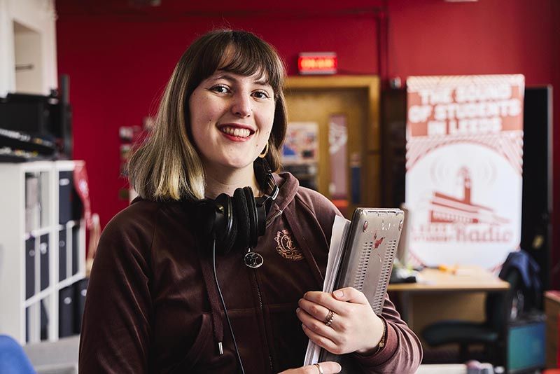 Carys gwenllian hind at leeds student radio. She is smiling and holding a laptop.
