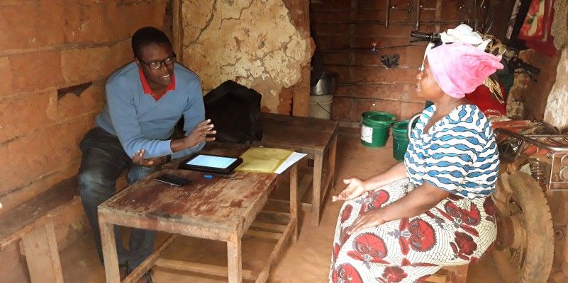 Two people sitting and talking in a room, with buckets and a motorbike behind them. One person has a tablet and phone in front of them on a table.